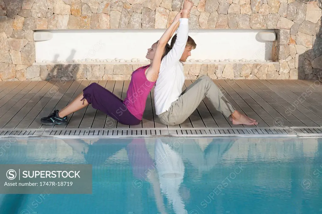 Young couple sitting back to back at poolside stretching
