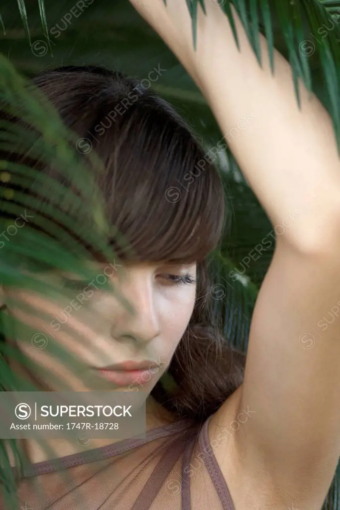 Young woman behind palm frond, looking down, portrait