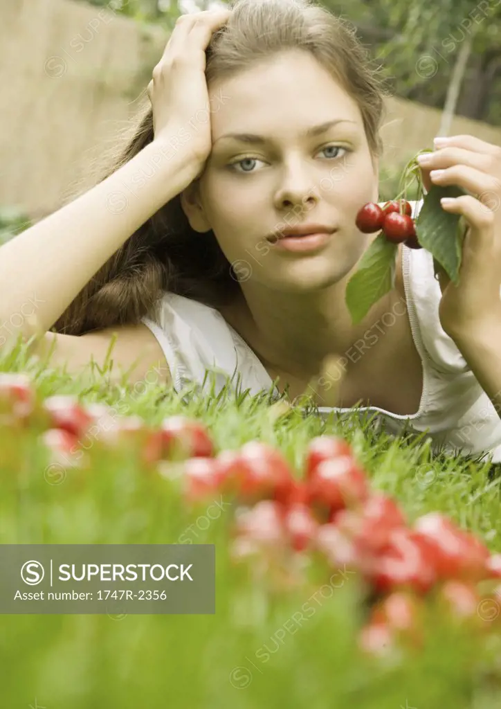 Woman lying in grass holding up cherries, looking at camera