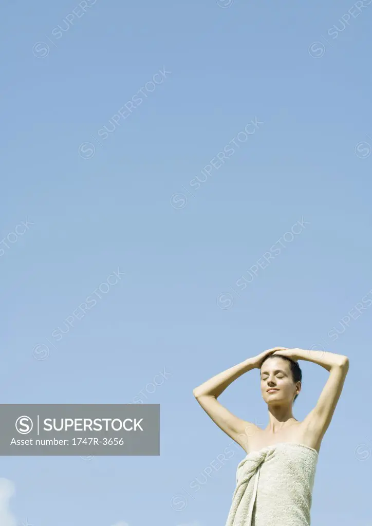 Woman standing wrapped in towel, hands on head, sky in background