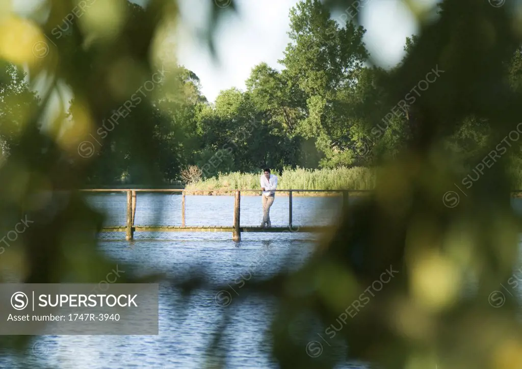 Man standing on footbridge over river, in distance, seen through leaves in blurred foreground