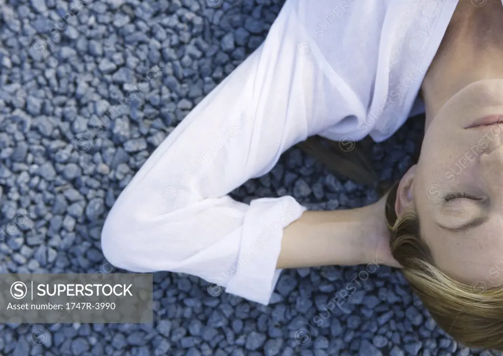 Woman lying on gravel with eyes closed and hands behind head, cropped