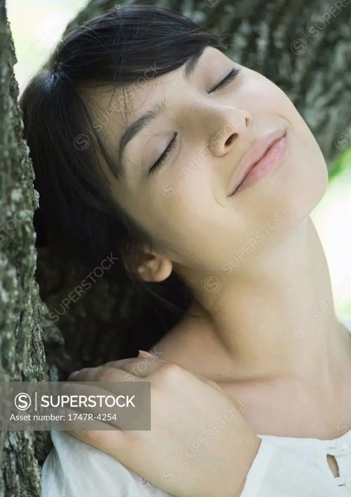 Woman with head against tree trunk, eyes closed, smiling