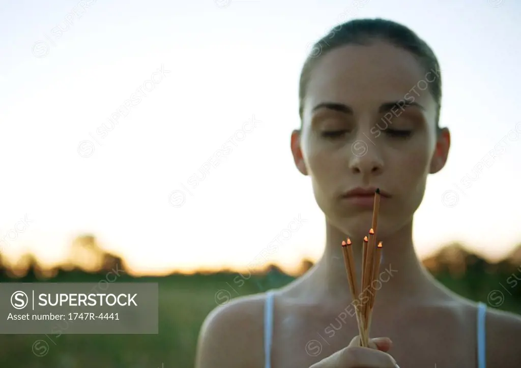 Woman holding bunch of incense, eyes closed, at sunset