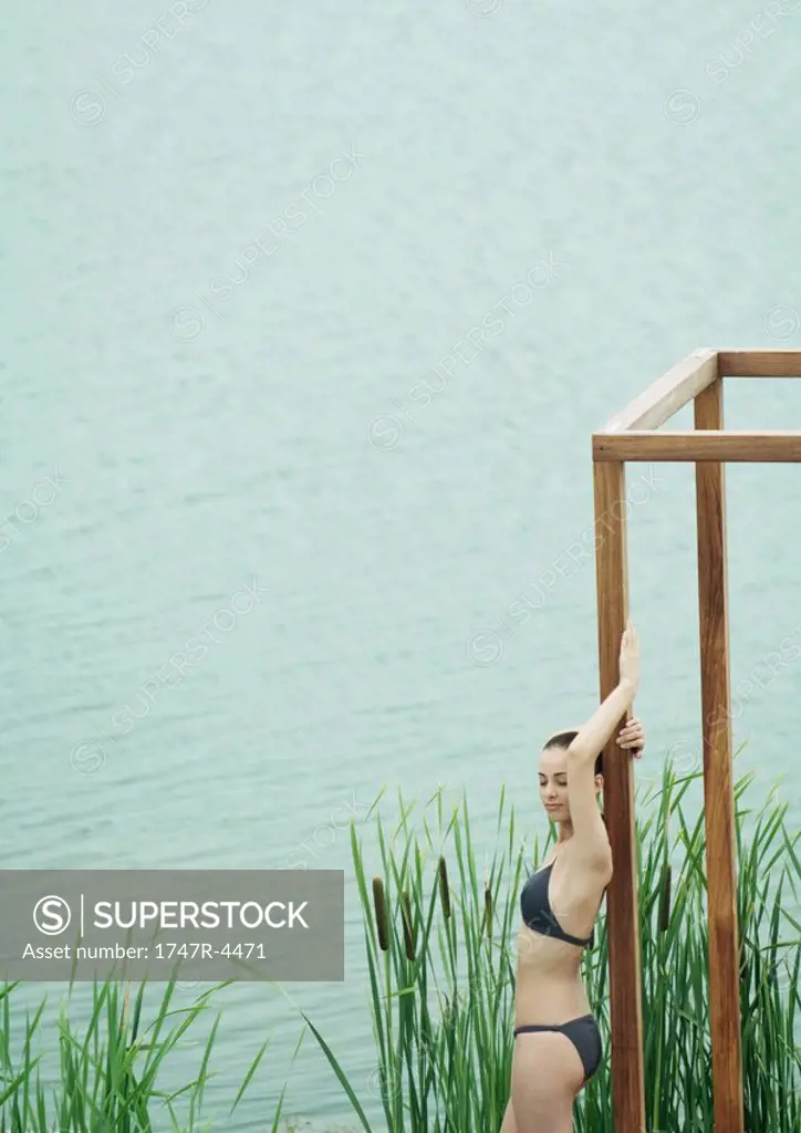 Woman wearing bikini, leaning against wooden structure, near lake