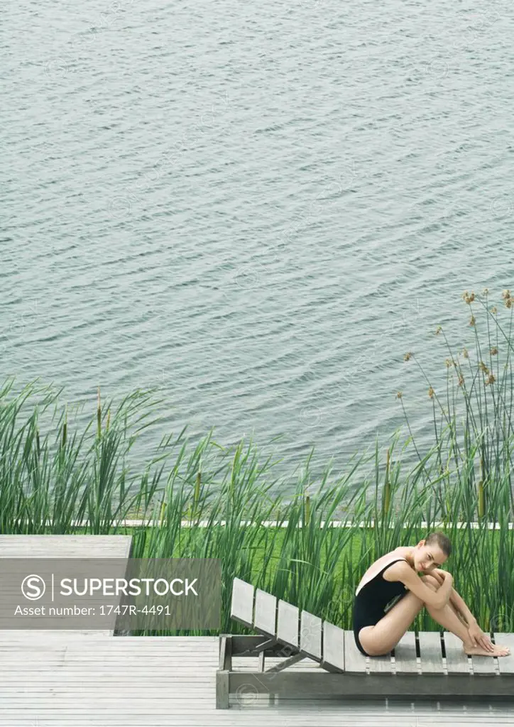 Woman sitting on lounge chair next to lake, hugging knees, smiling
