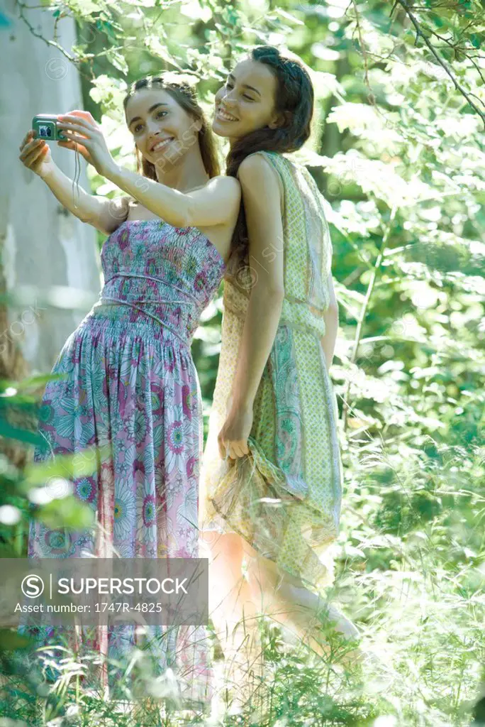 Two young women wearing sundresses, standing in forest, taking photo of selves with digital camera