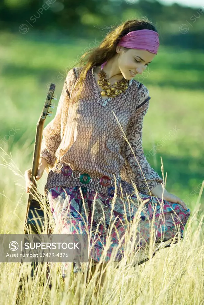Young hippie woman walking through field, holding guitar
