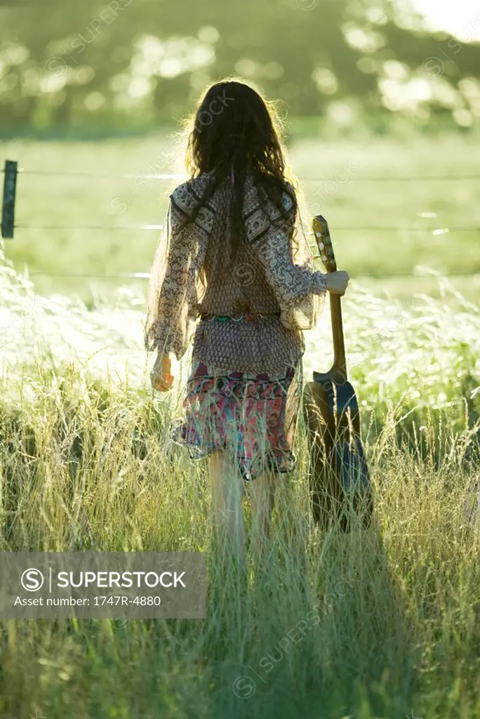 Young hippie woman holding guitar, standing in field, rear view