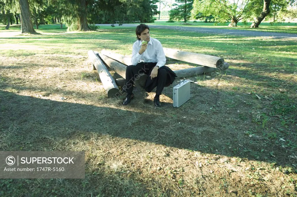 Businessman sitting on logs, eating an apple, looking away
