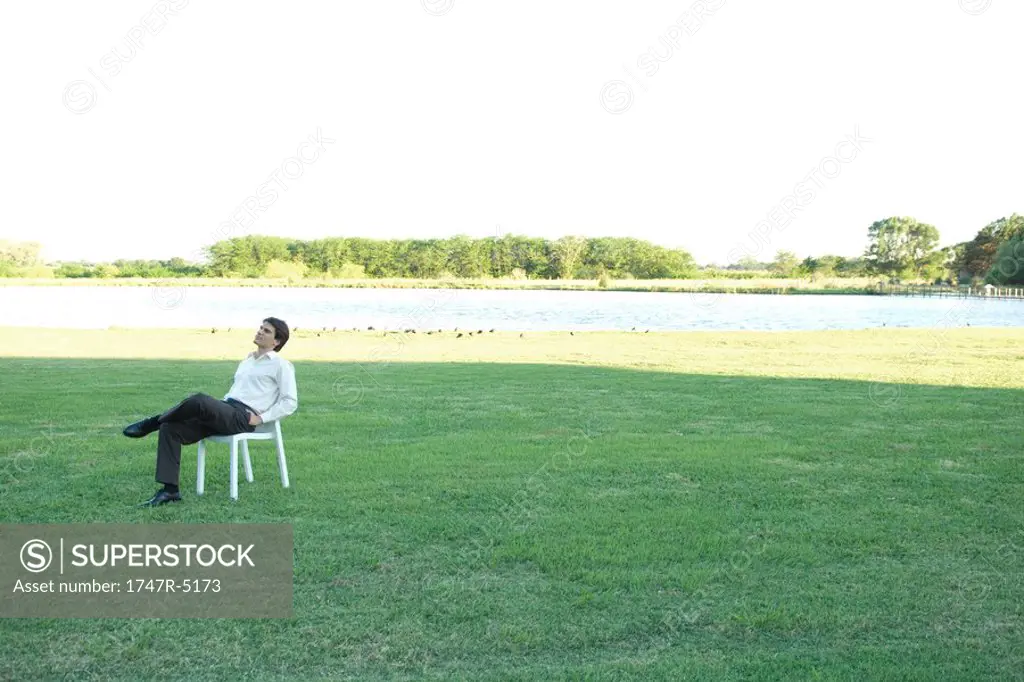 Businessman sitting in chair on lawn, hands in pockets, looking up