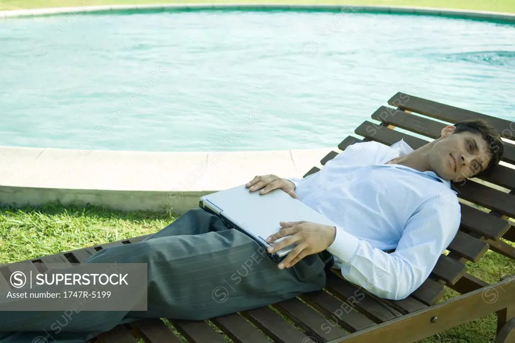 Businessman reclining in lounge chair near pool, holding laptop on lap
