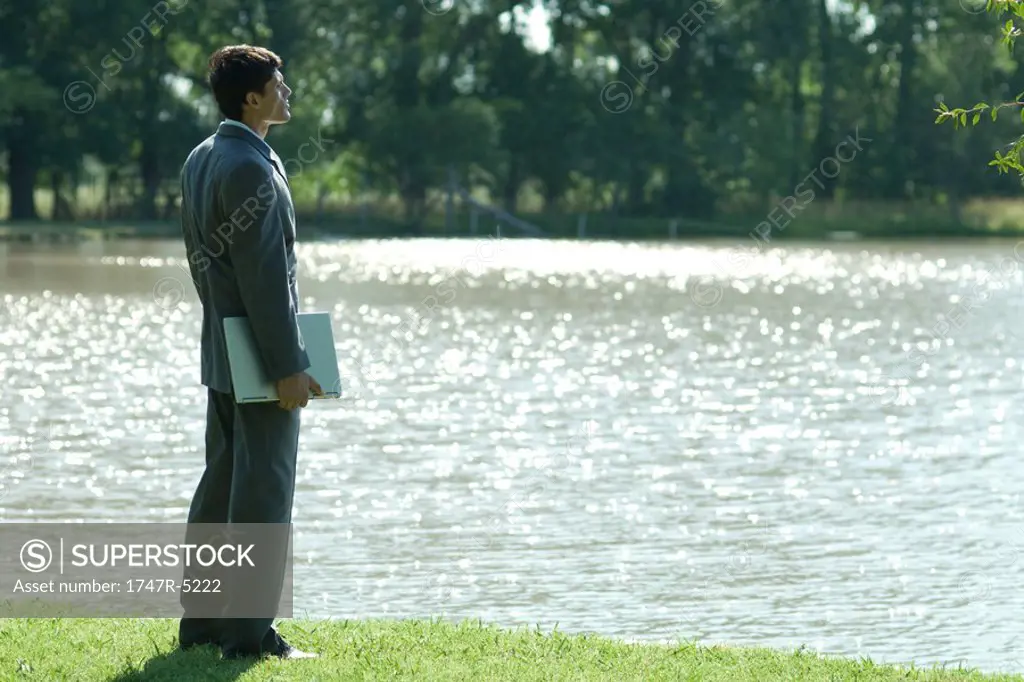 Businessman standing by edge of water, holding laptop under arm