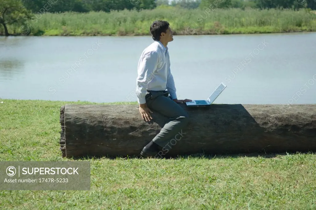 Businessman sitting on tree trunk with laptop, looking away
