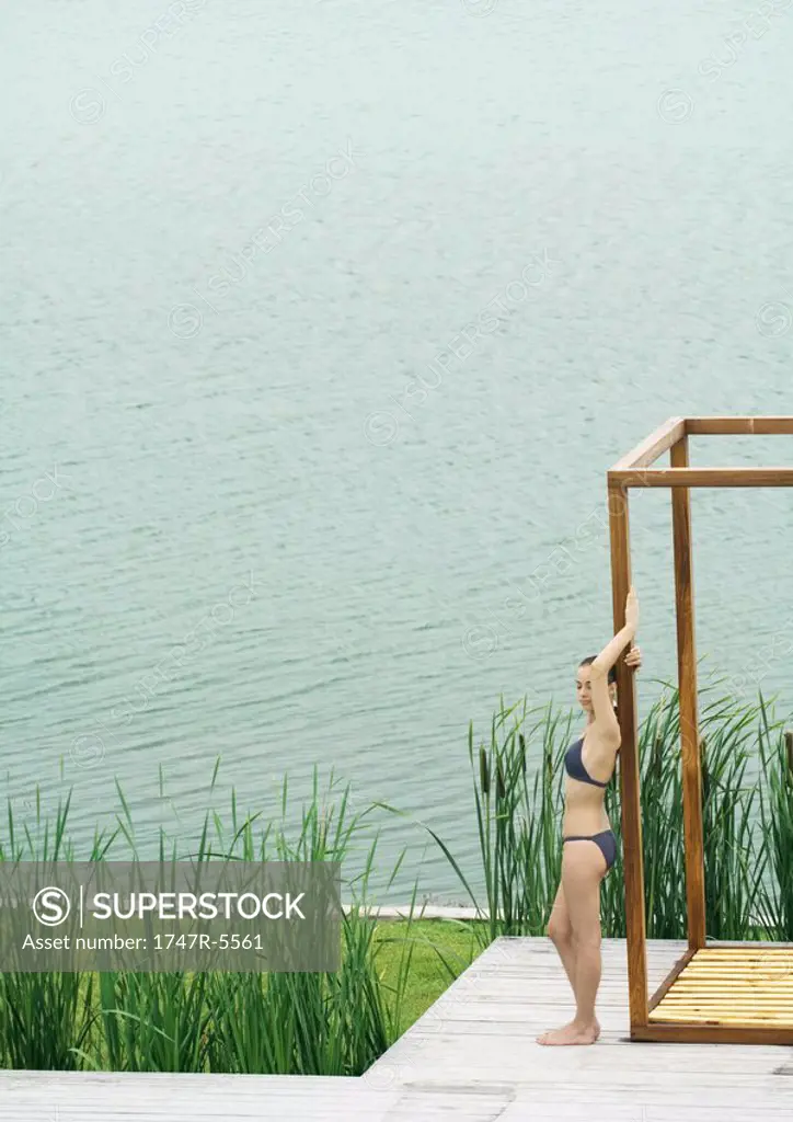 Young woman in swimsuit standing by edge of water, leaning against wooden structure