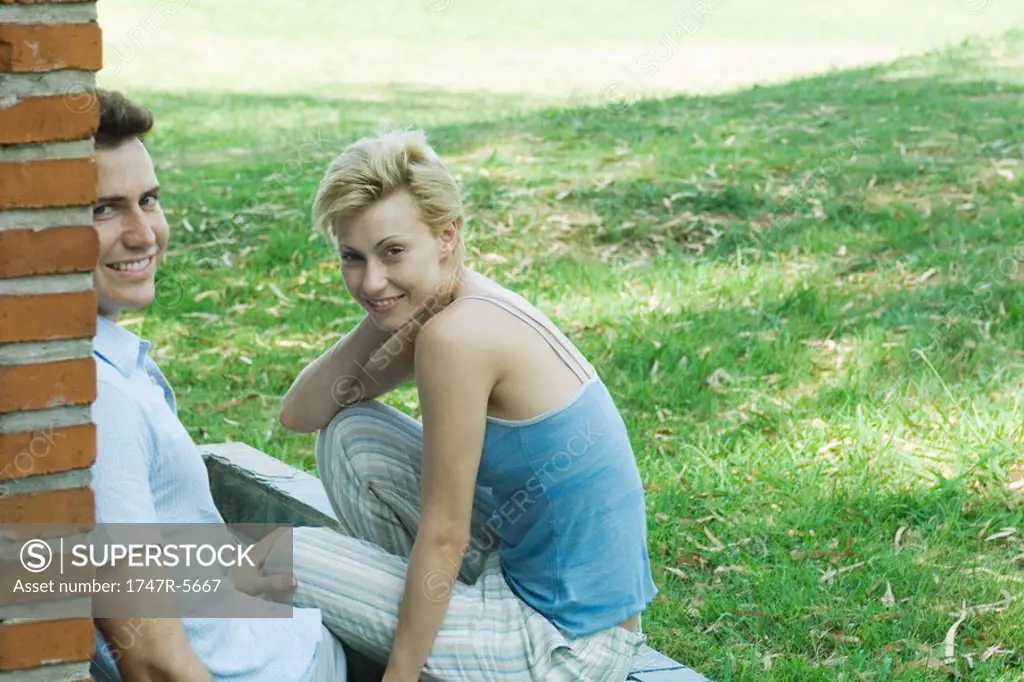 Couple sitting outdoors, next to brick wall, smiling at camera