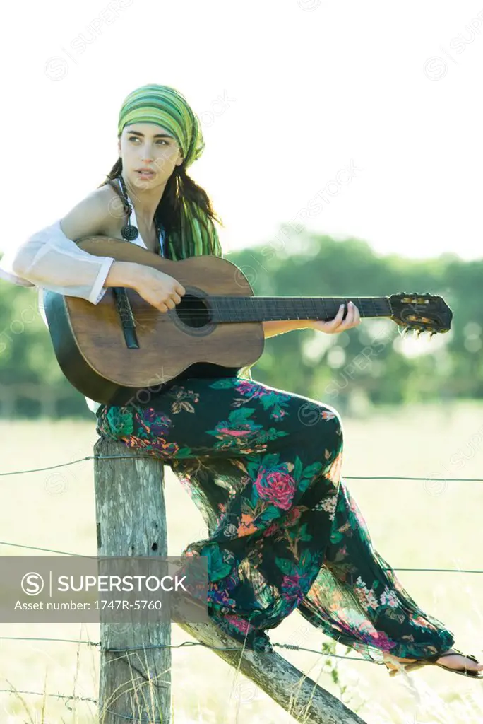 Young hippie woman sitting on fence post, playing guitar