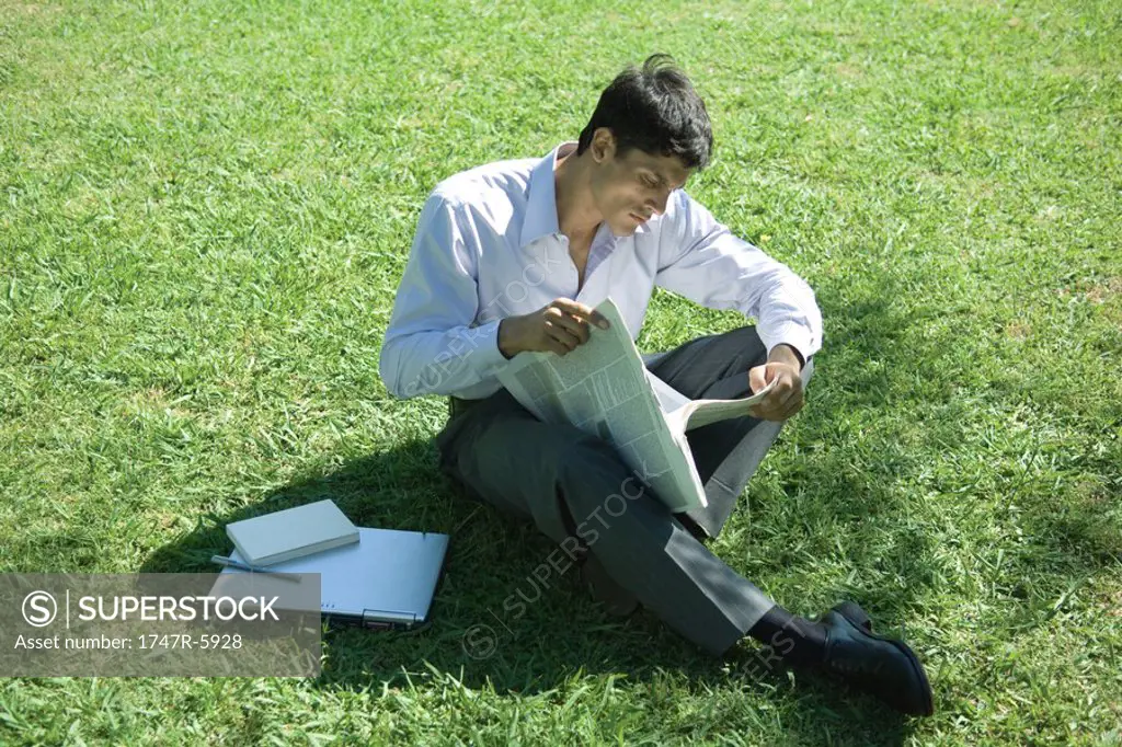Businessman sitting on grass, reading newspaper