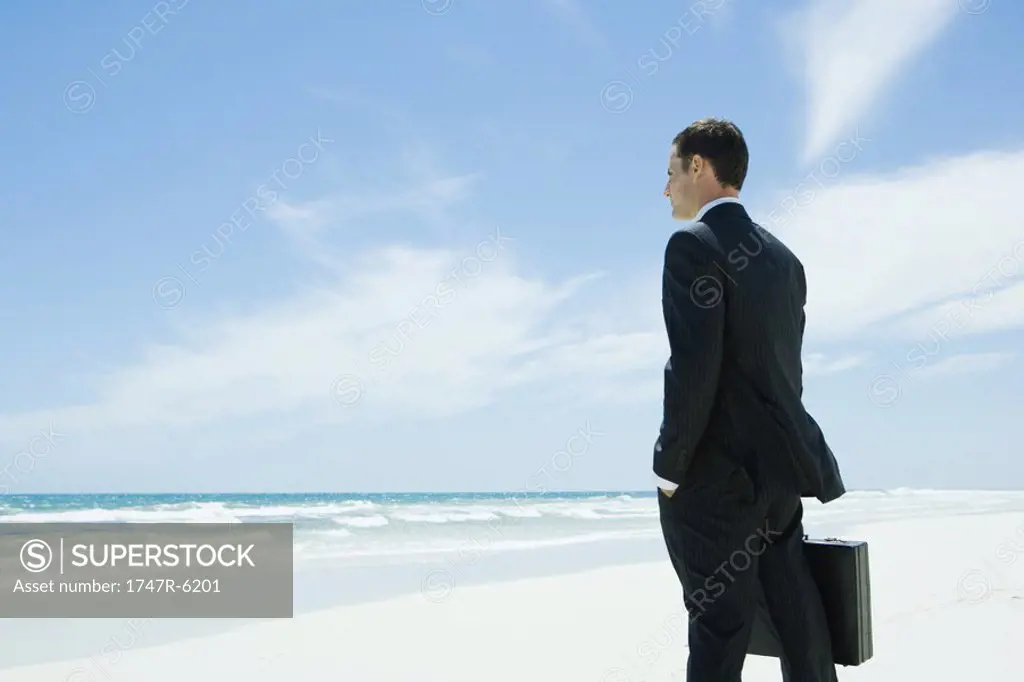 Businessman standing on beach, holding briefcase, facing ocean