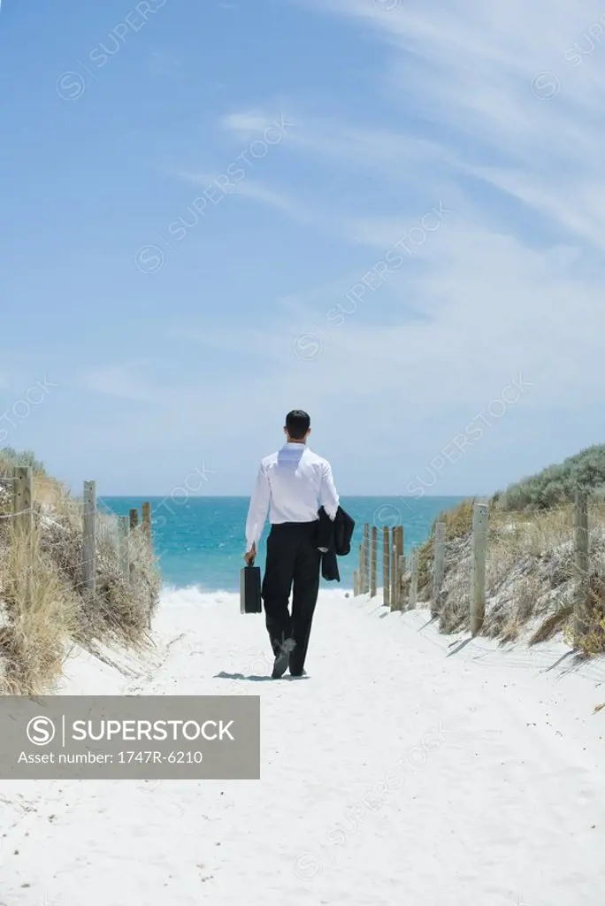 Businessman walking toward beach, holding briefcase and jacket over arm, rear view