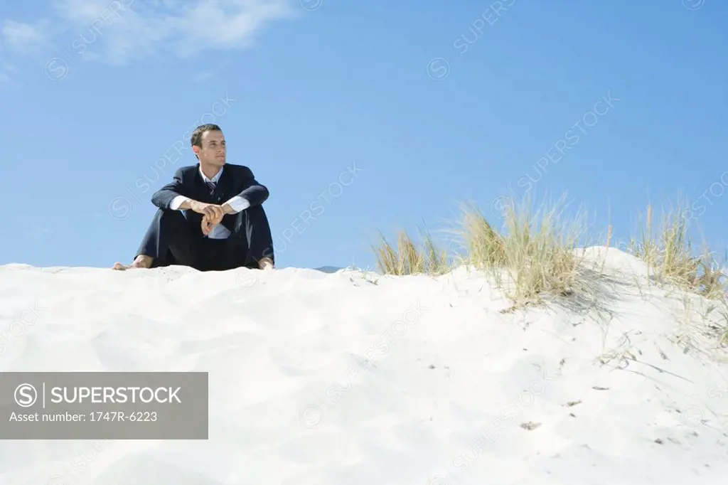 Businessman sitting on dune, looking at distance