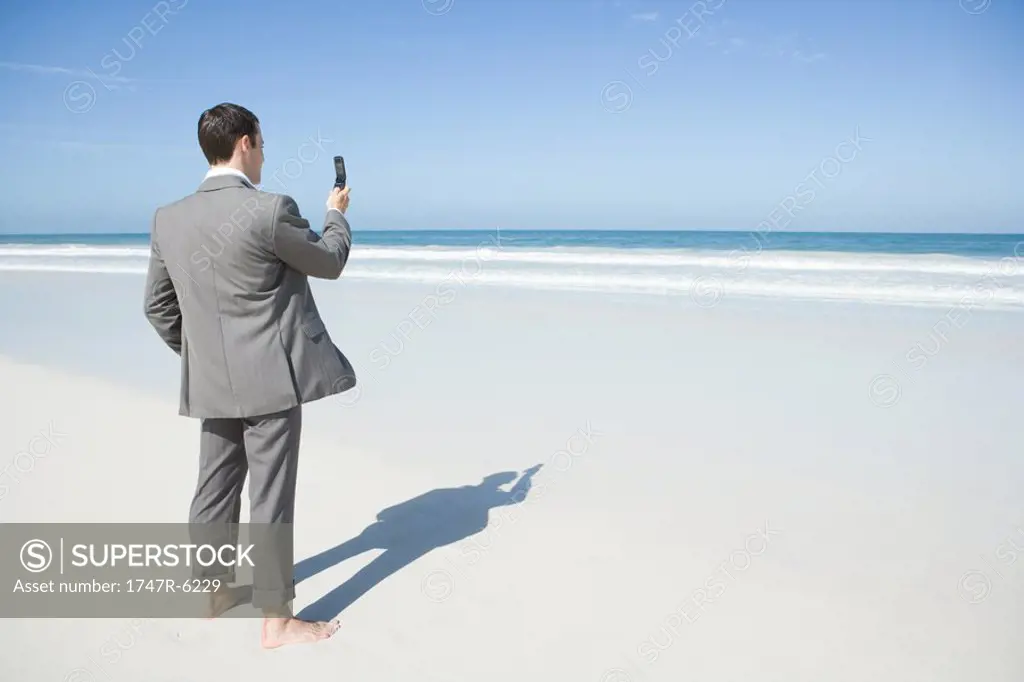 Barefoot businessman standing on beach, holding up cell phone