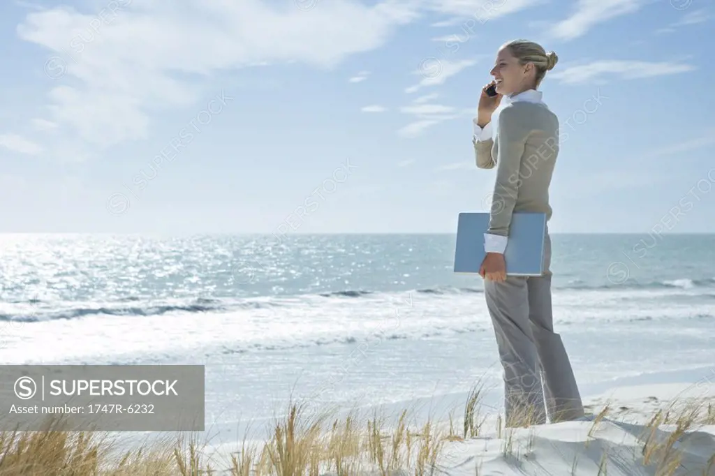 Businesswoman standing on beach, holding laptop and using cell phone