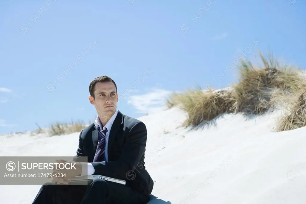 Businessman sitting on beach, holding laptop on lap, looking away