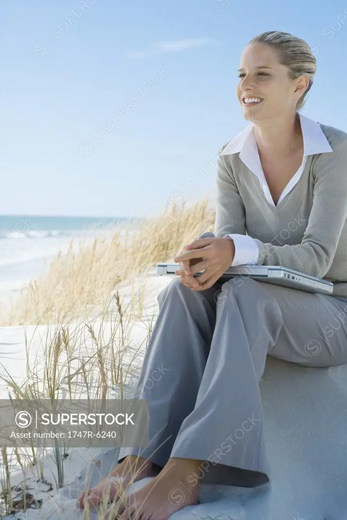 Barefoot businesswoman sitting on dune, laptop on lap, smiling