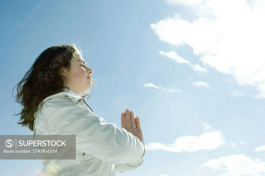 Teen girl standing with hands in prayer position and eyes closed, low angle view