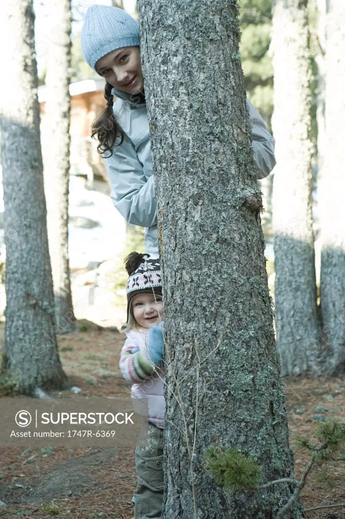Teen girl and toddler, peeking out from behind tree trunk