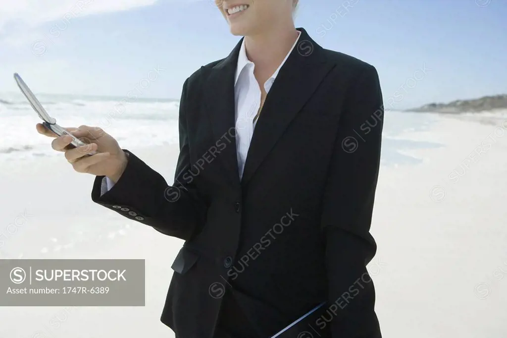 Businesswoman holding cell phone, standing on beach, smiling, cropped