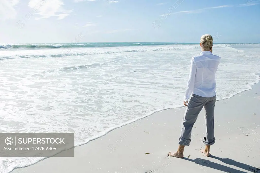 Woman standing barefoot on beach, using cell phone, rear view