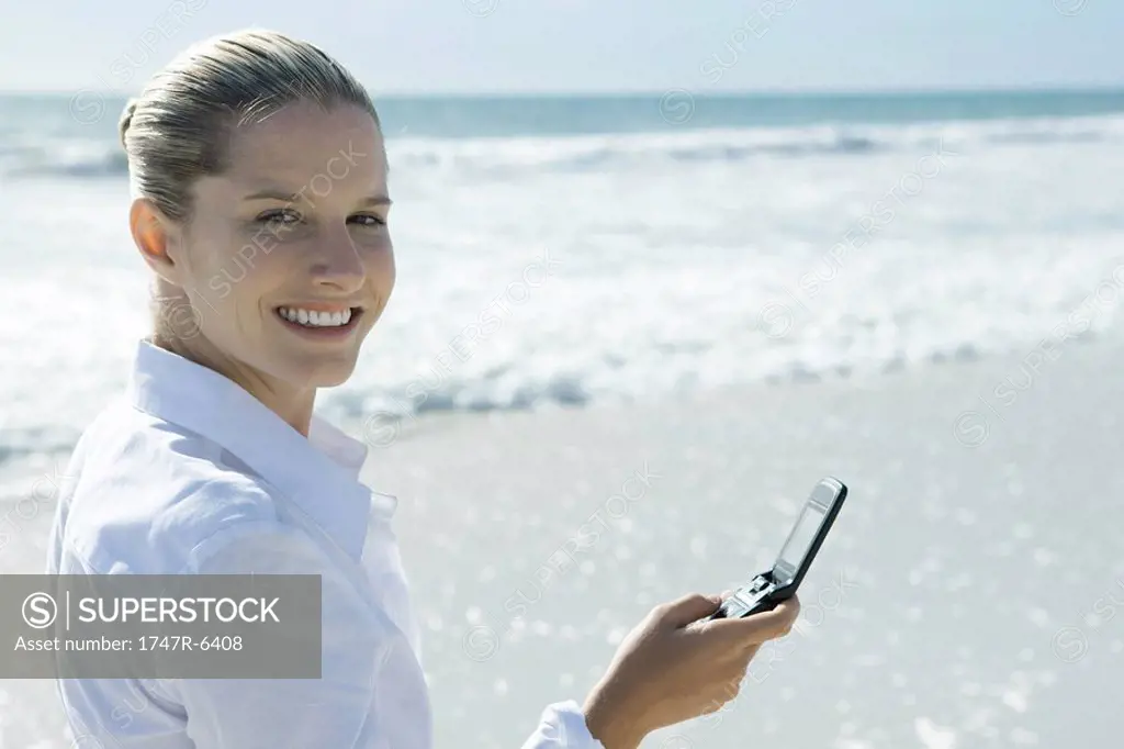 Woman on beach, holding up cell phone, looking over shoulder at camera