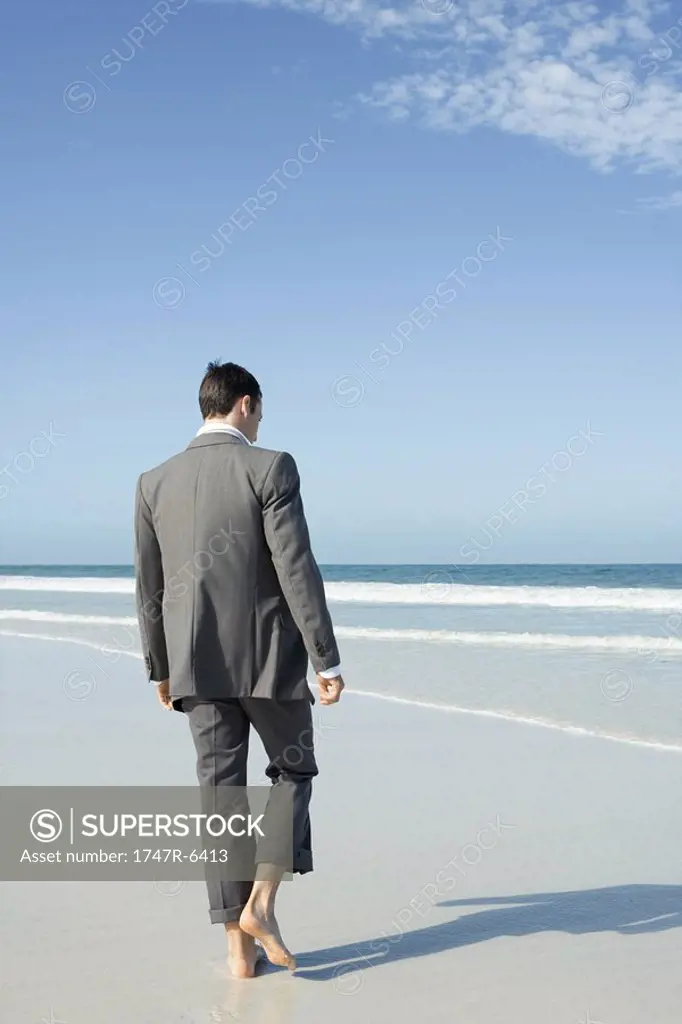 Businessman walking barefoot on beach, rear view