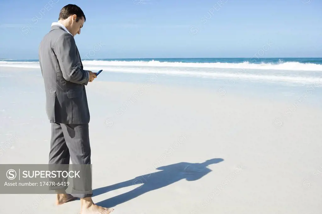 Businessman standing barefoot on beach, looking at cell phone