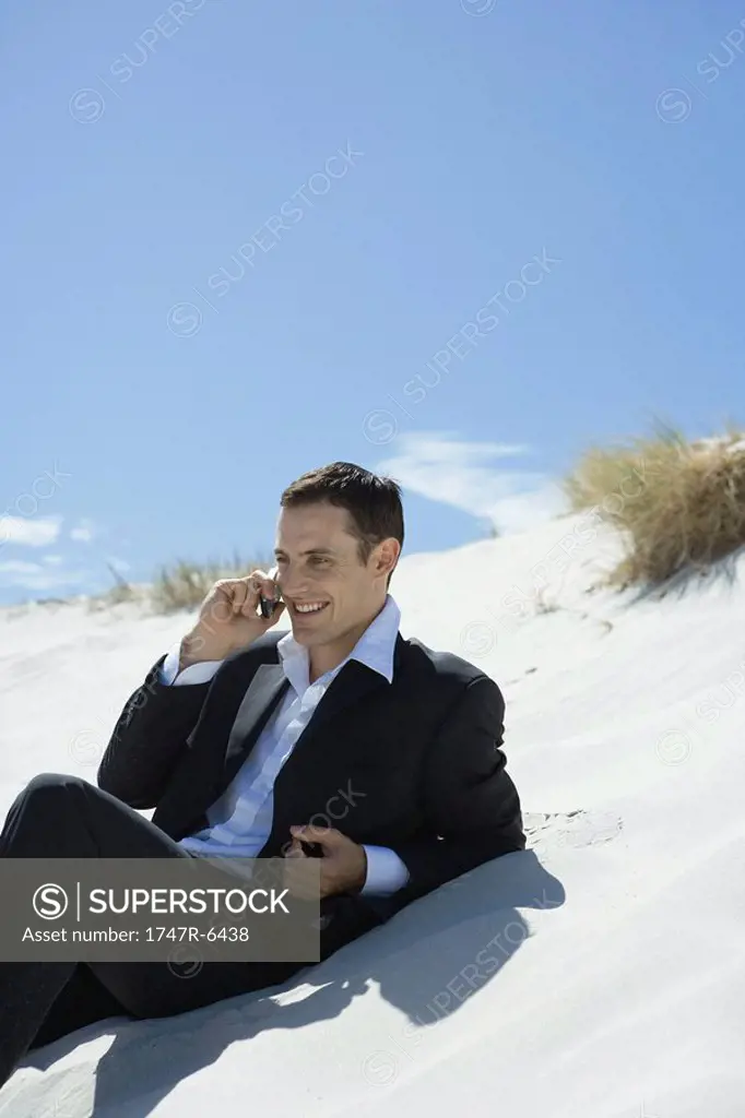 Businessman sitting on sand dune, using cell phone