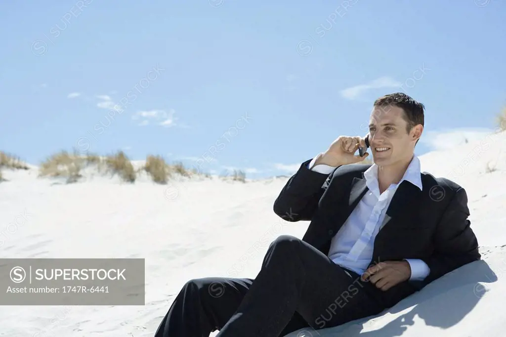 Businessman sitting on sand dune, using cell phone