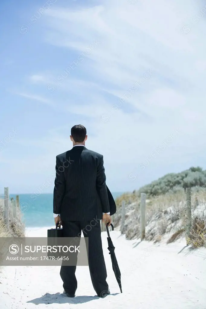 Businessman standing on sandy path leading to ocean, holding briefcase and umbrella, rear view