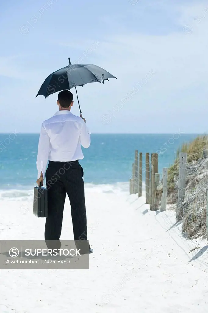 Businessman standing under umbrella on sandy path leading to ocean, rear view