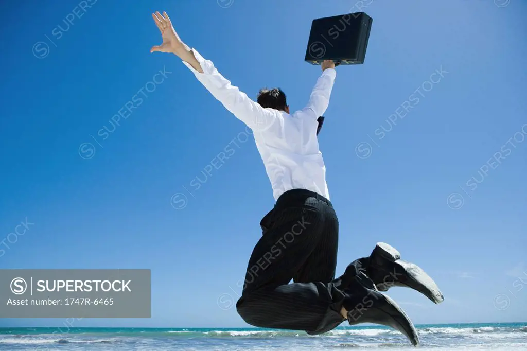 Businessman on beach, jumping into air with briefcase in hand