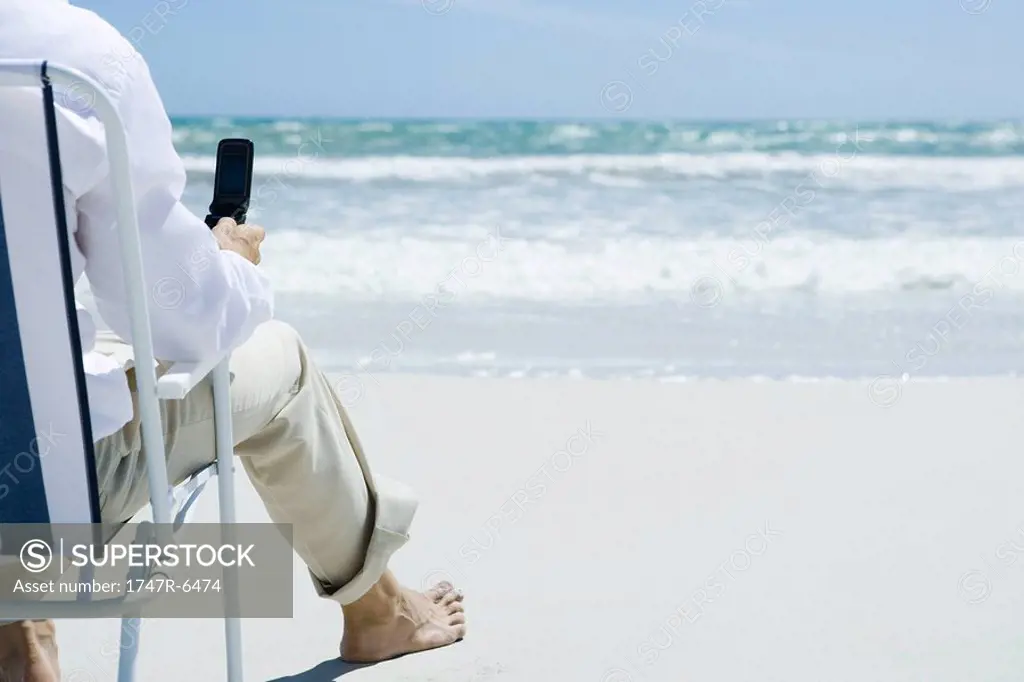 Man sitting in folding chair on beach, holding cell phone, cropped, rear view