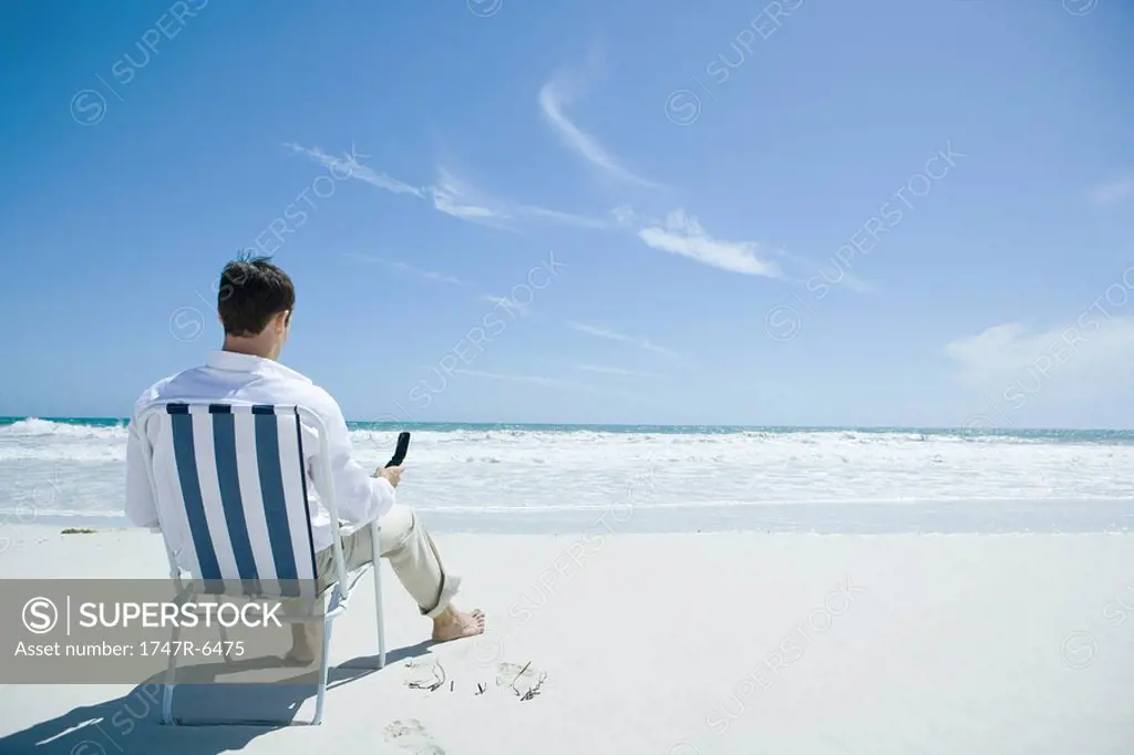 Man sitting in folding chair on beach, holding cell phone, rear view