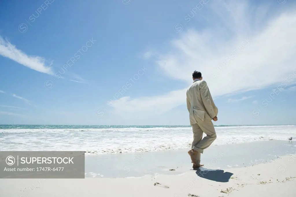 Businessman walking barefoot on beach, rear view