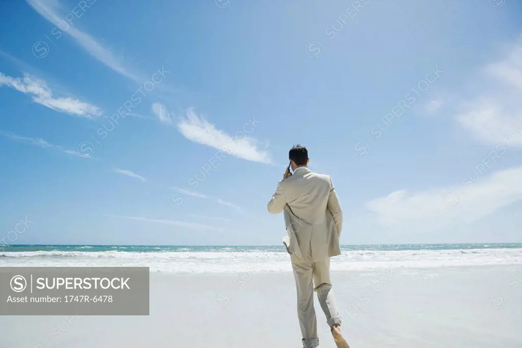 Businessman walking barefoot on beach, using cell phone