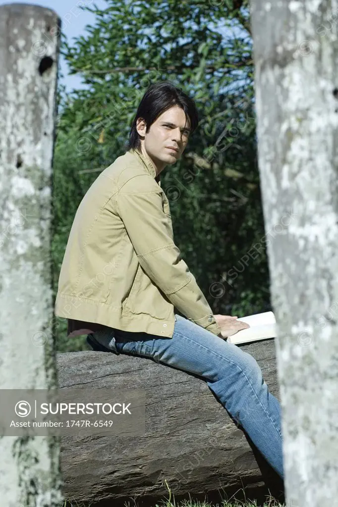 Man sitting on tree trunk with book, looking away