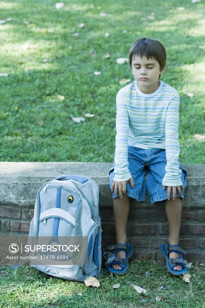 Boy sitting on low wall, backpack by side, eyes closed