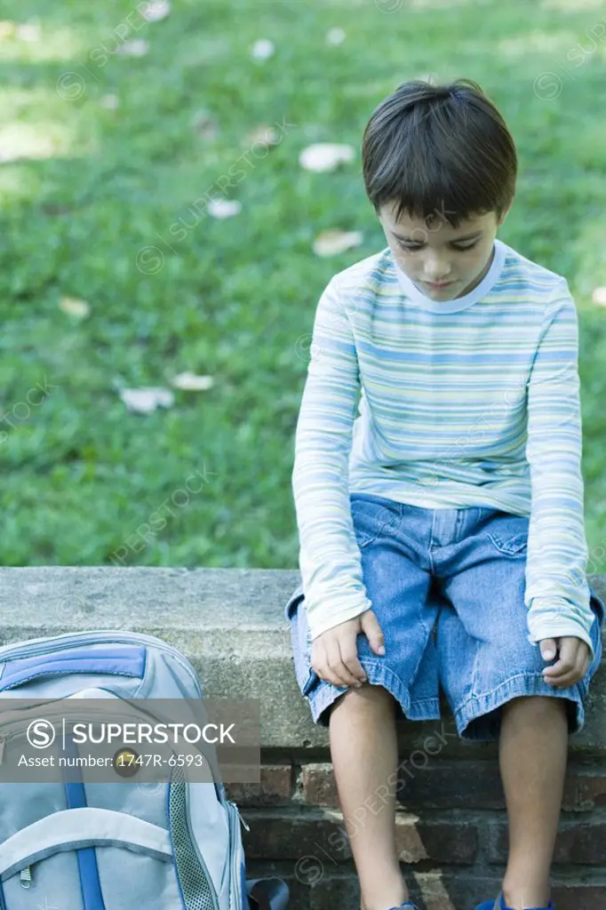 Boy sitting on low wall, backpack by side, looking down