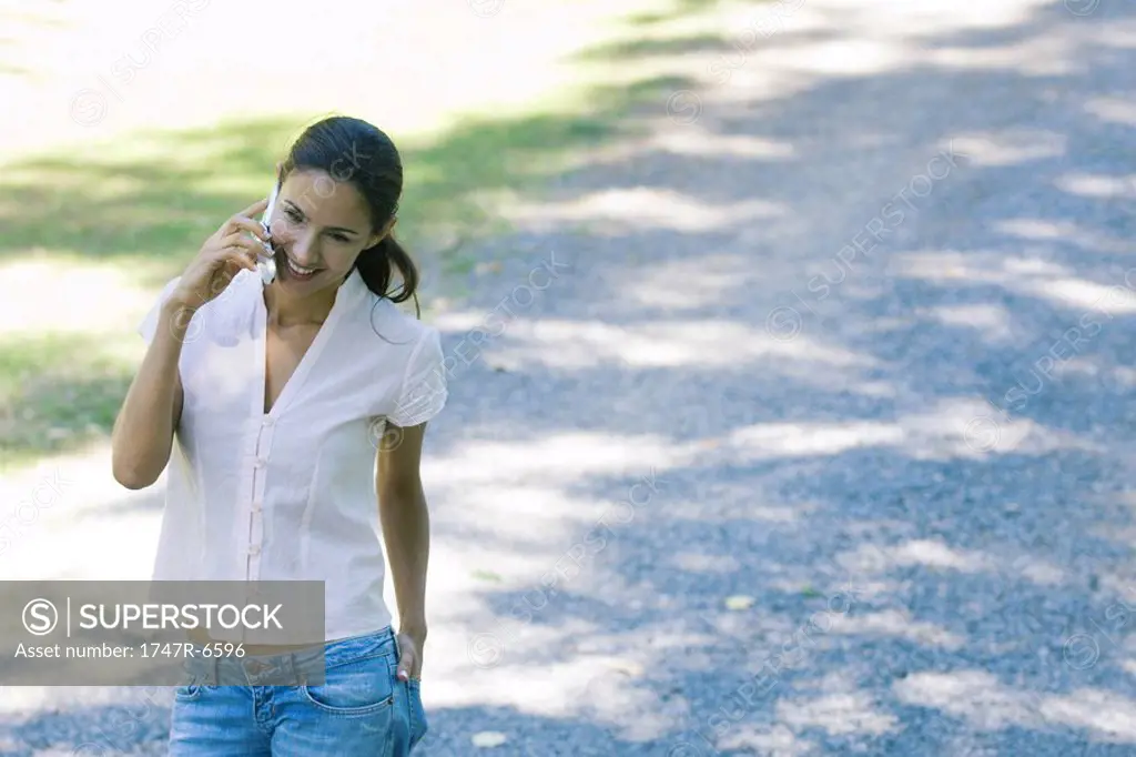 Casually dressed woman using cell phone, smiling as she walks through park