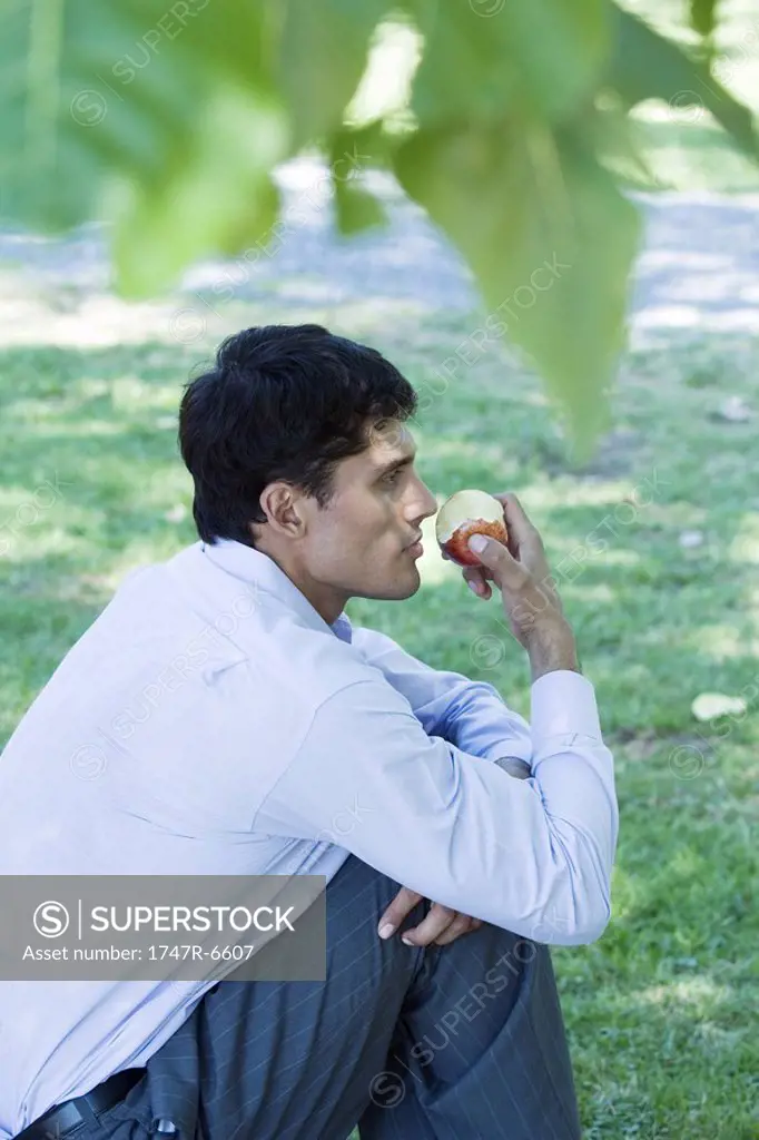 Businessman sitting on grass, eating apple, side view